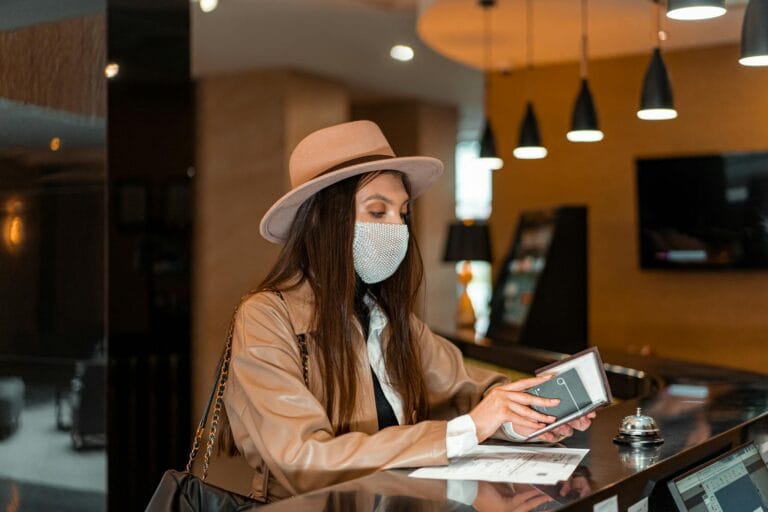 Woman wearing mask and hat checking in at hotel front desk, symbolizing travel and safety.
